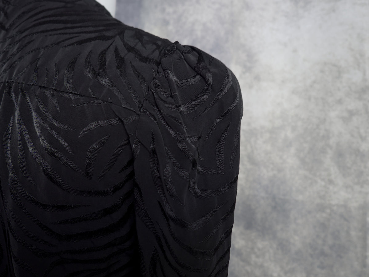 Close-up of a black textured garment with a zebra-like jacquard pattern, showing the shoulder and upper back against a mottled gray background.