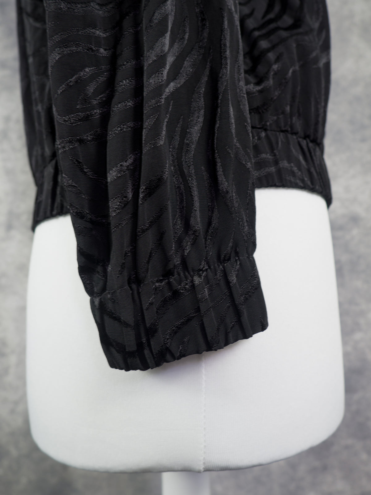 Black long-sleeve blouse with a subtle wavy jacquard pattern and gathered elastic cuffs, displayed on a white mannequin torso against a gray background.