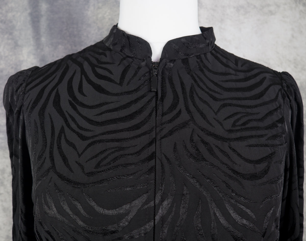 Black long-sleeve blouse with a textured zebra-stripe jacquard pattern, featuring a mock neck and front zipper, displayed on a white mannequin torso.