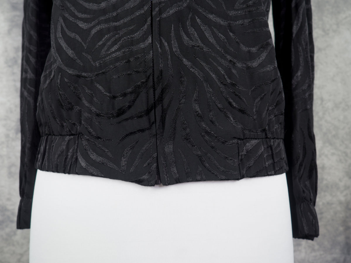 A black long-sleeve cropped jacket with a textured zebra-like jacquard pattern, displayed on a white mannequin torso against a gray textured background.