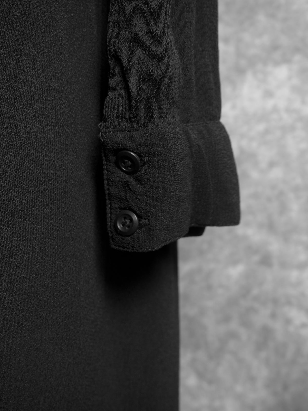 A close-up shot of the sleeve cuff on a black dress, showcasing two small black buttons on a classic buttoned cuff. The image highlights the soft, lightweight texture of the fabric and the neat stitching details, set against a blurred grey background.