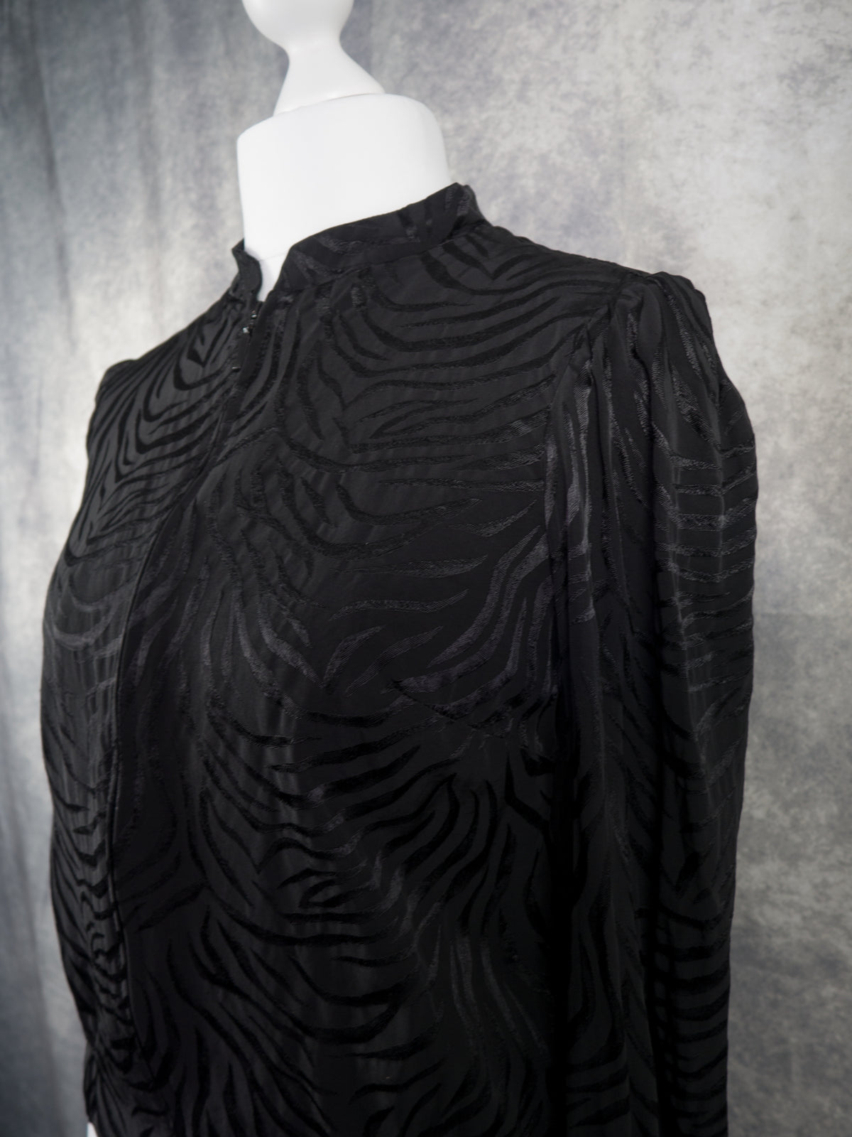 A black long-sleeve blouse with a textured, wavy jacquard pattern, displayed on a white mannequin torso against a gray textured backdrop.
