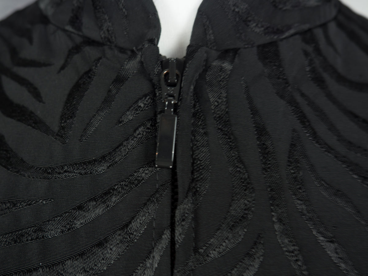 Close-up of a black textured zip-up garment with a subtle wavy jacquard pattern, featuring a black zipper pull and a white mannequin neck visible at the top.