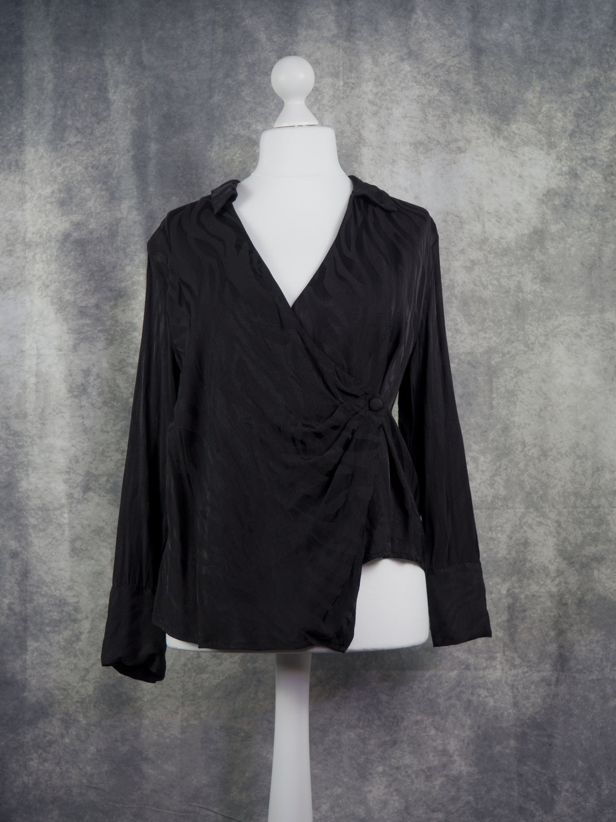 A black long-sleeve wrap-style blouse displayed on a white mannequin torso against a textured gray studio backdrop.