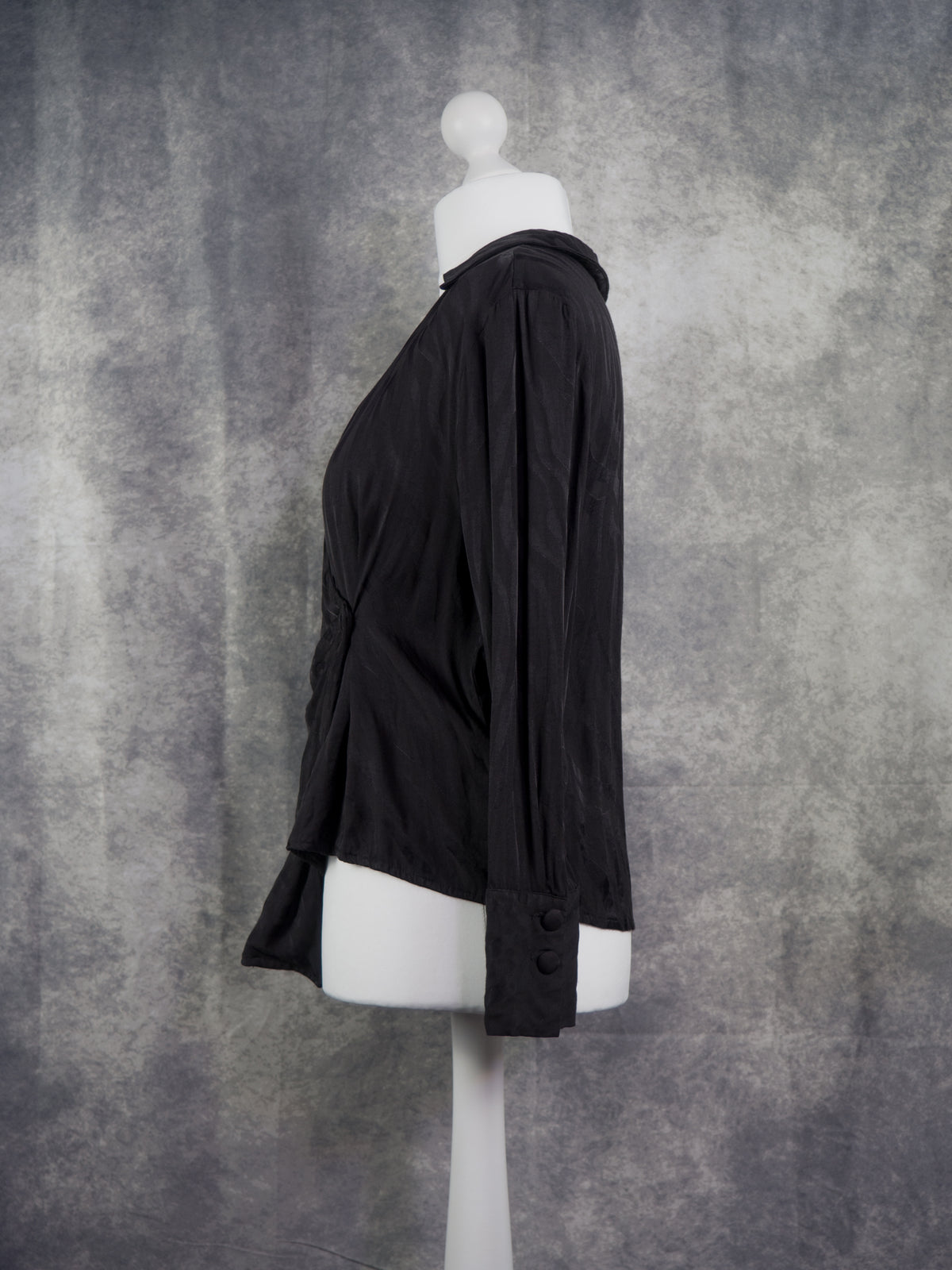 A black long-sleeve draped blouse displayed on a white mannequin torso against a textured gray studio backdrop.