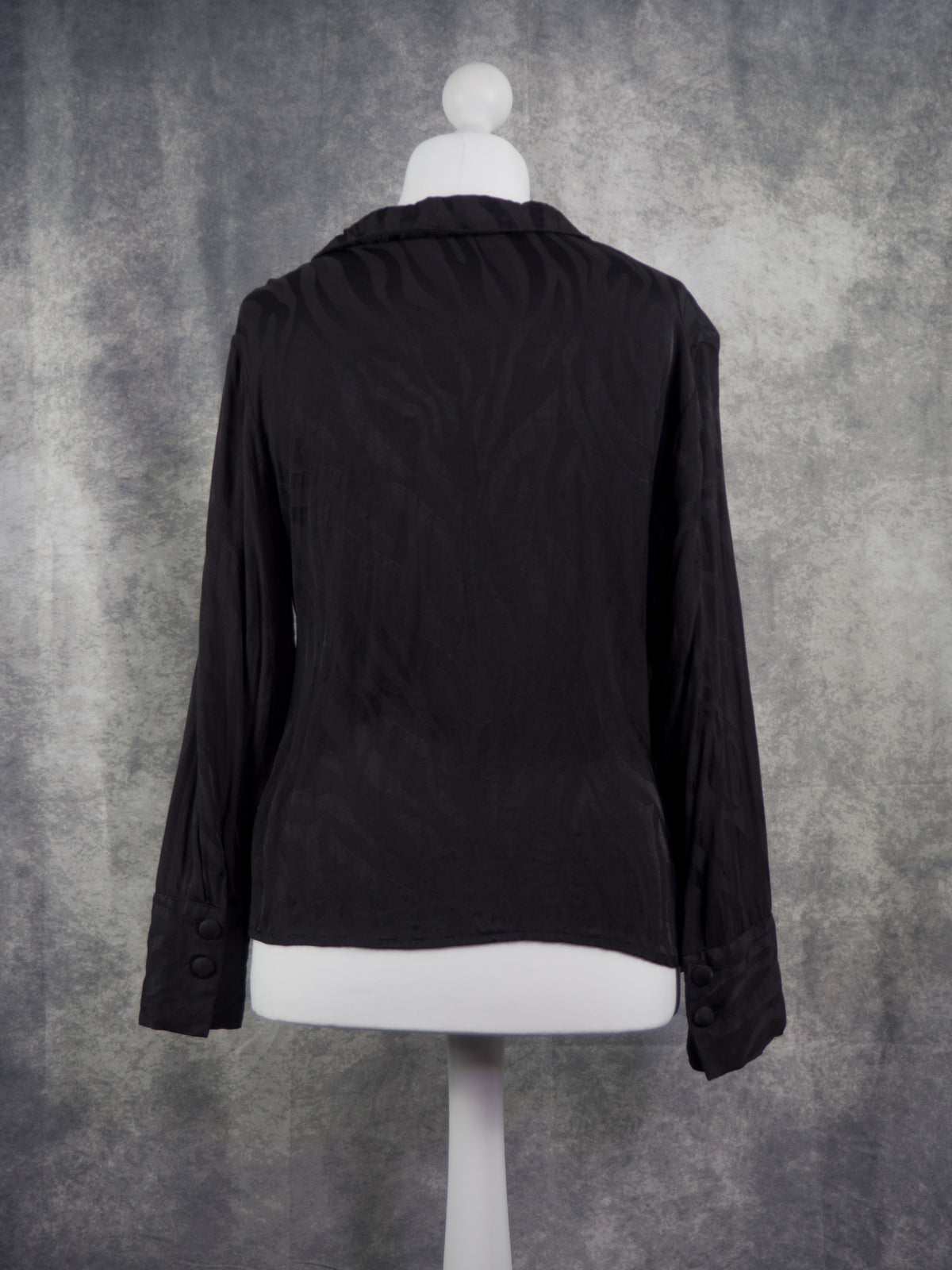 A black long-sleeve blazer shown from the back on a white mannequin torso against a textured gray studio backdrop.