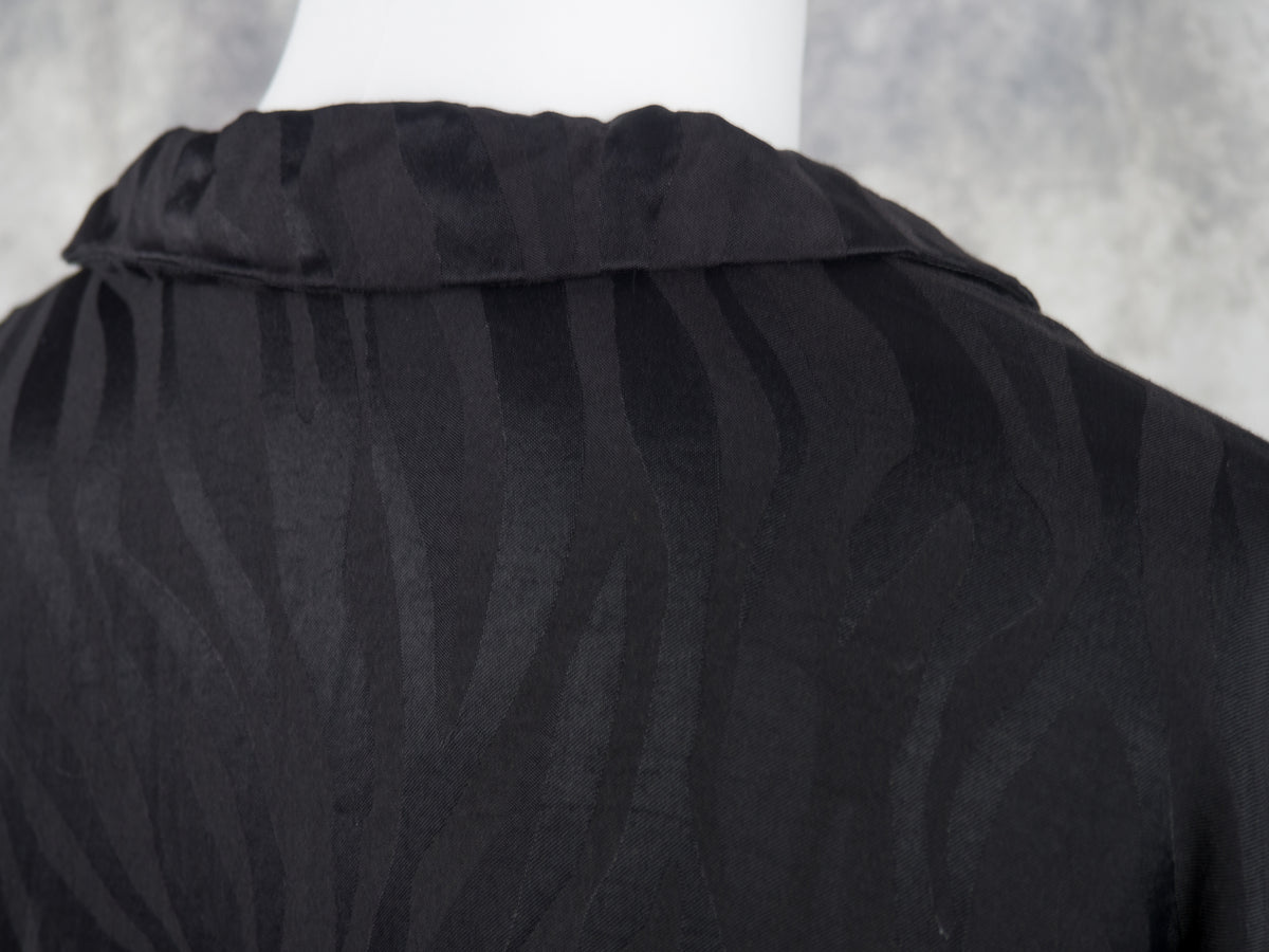 Close-up of a black textured blouse or top displayed on a white mannequin torso, showing a subtle wavy jacquard pattern and collar detail against a soft gray background.
