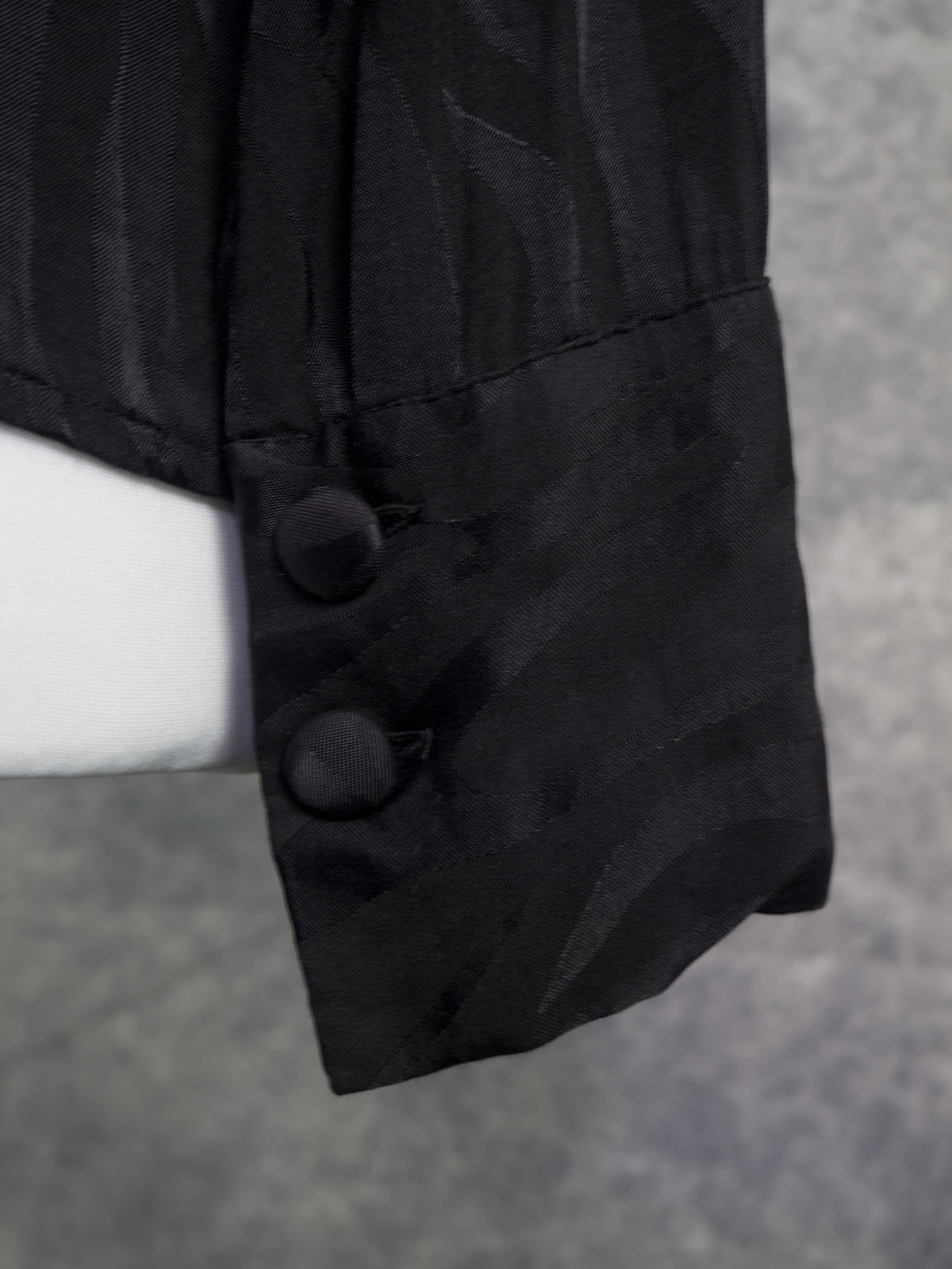 Close-up of a black textured fabric sleeve or cuff with two dark buttons, shown against a gray background with a white mannequin support visible on the left.