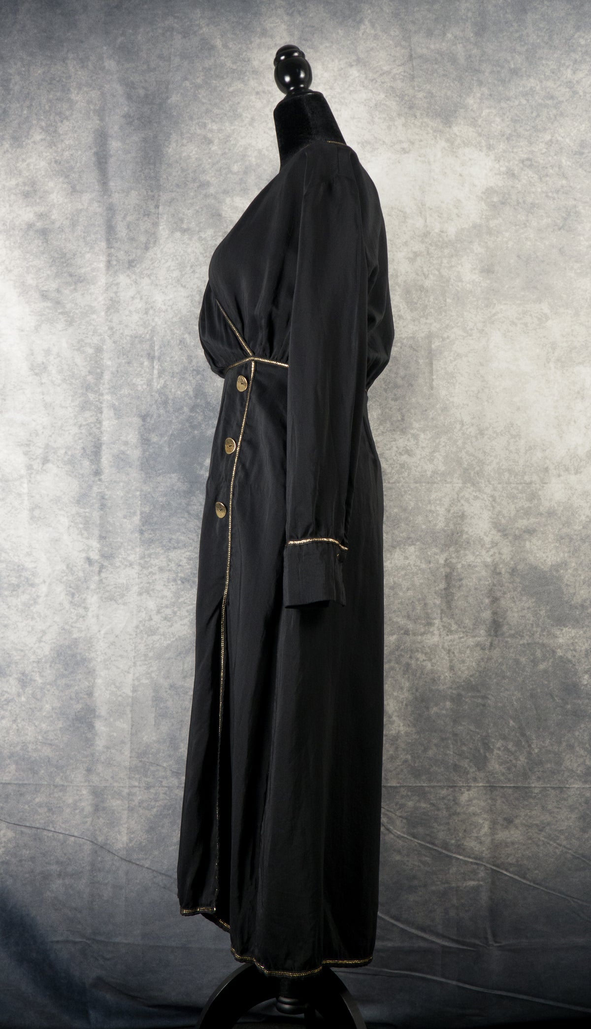 A full-length side profile view of a black maxi wrap dress displayed on a black mannequin against a textured gray background. The long-sleeved dress features a gathered waist accented with a thin gold trim. Three decorative round gold buttons are visible vertically along the front wrap seam, which opens up into a subtle leg slit near the bottom hem.
