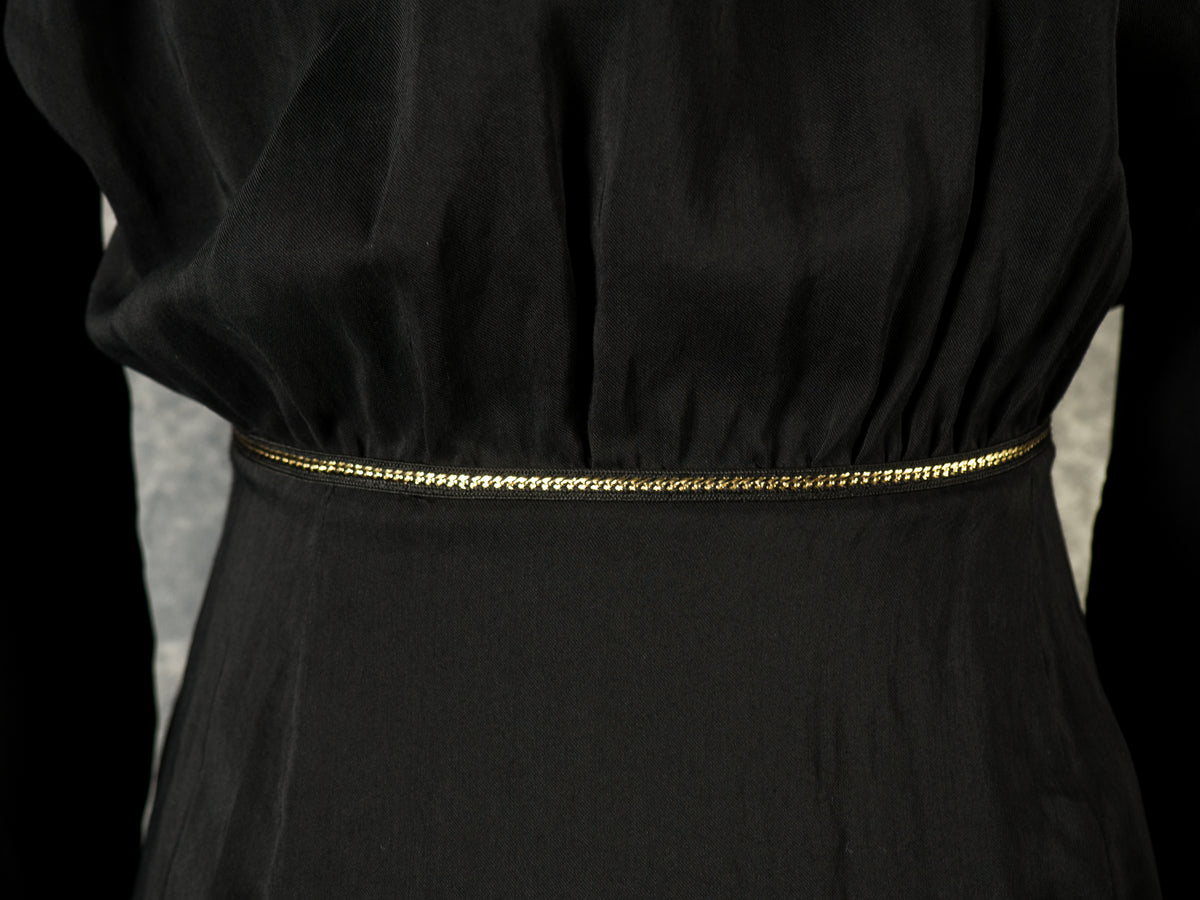 A close-up shot of the back waist section of a black maxi dress. The fabric of the bodice is softly gathered where it meets the straight-cut skirt at a fixed, non-elastic seam. A delicate metallic gold chain trim highlights the entire horizontal waistline against a dark backdrop.