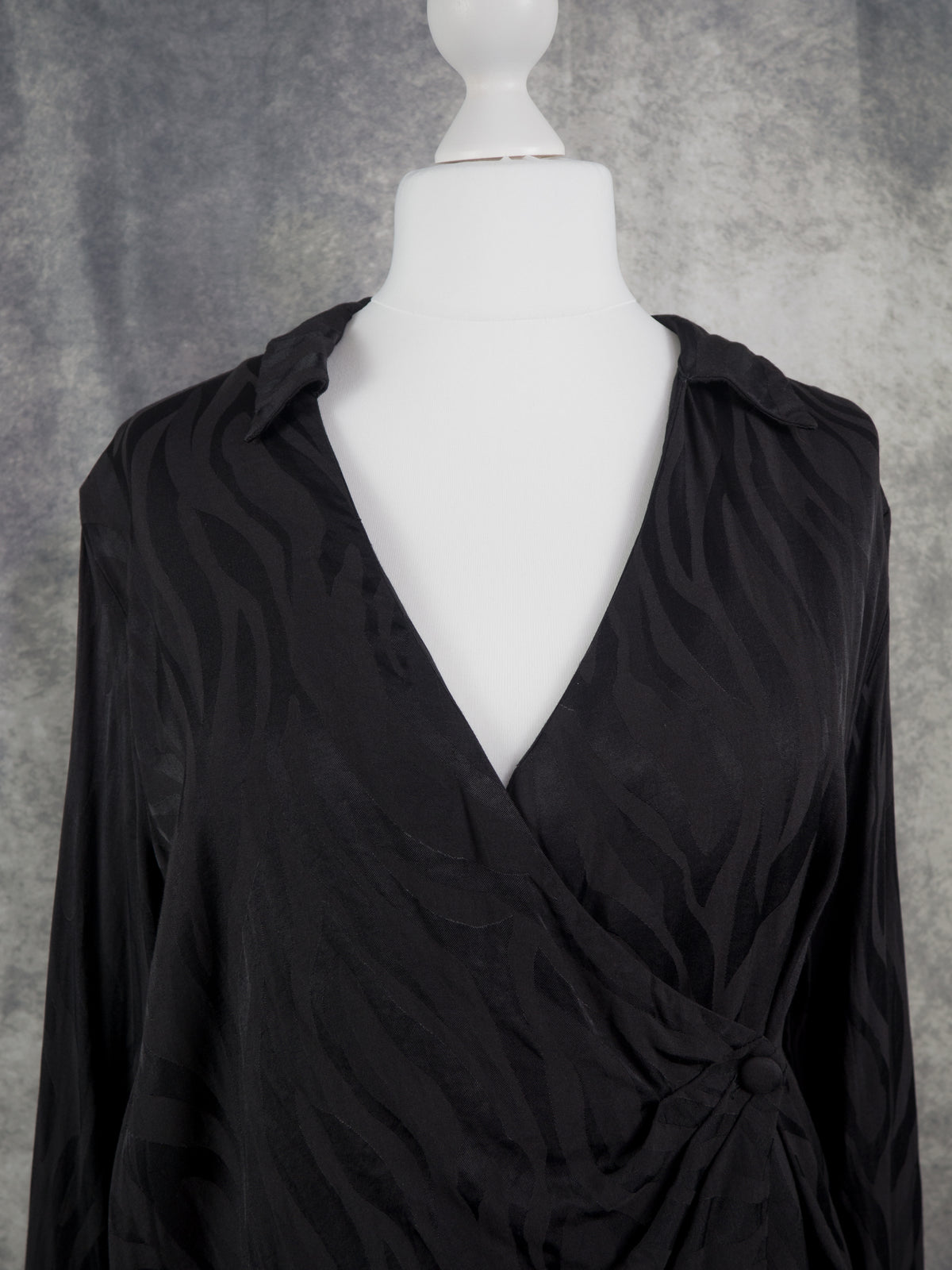 A black long-sleeve wrap-style blouse with a deep V-neck and gathered front detail, displayed on a white mannequin torso against a textured gray backdrop.