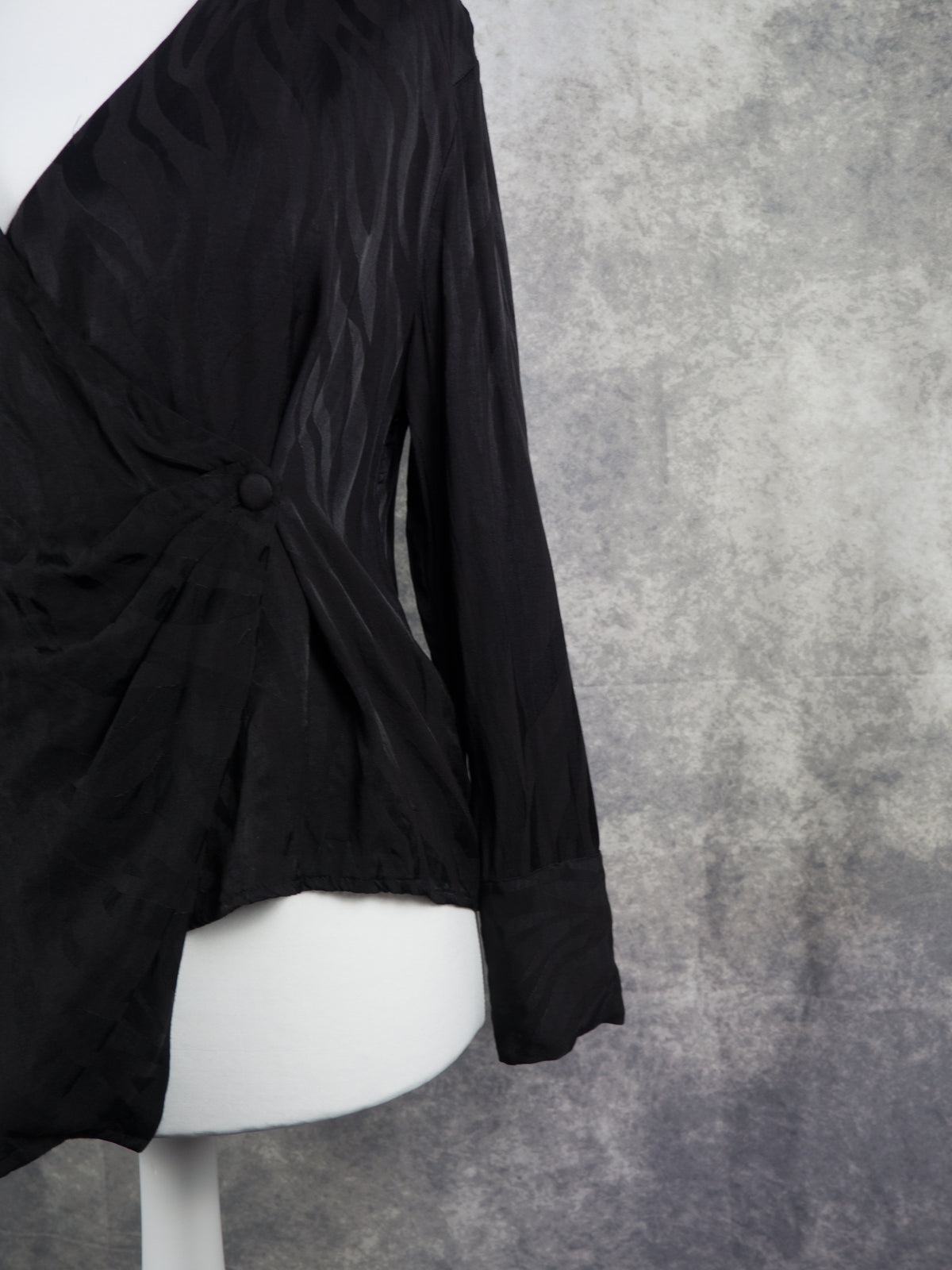 A black long-sleeve wrap-style blouse displayed on a white mannequin torso against a textured gray studio background.