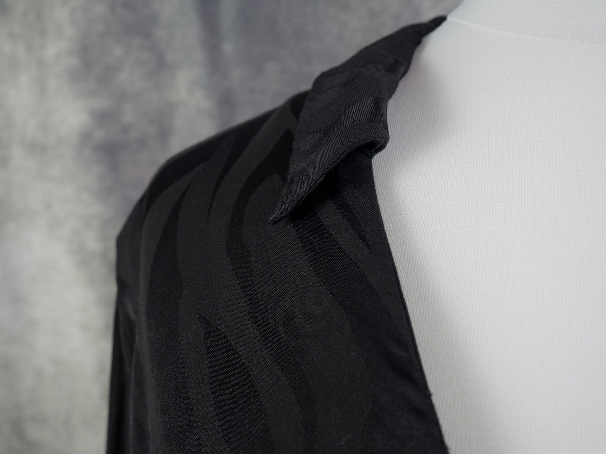 A close-up of a black long-sleeve collared shirt with a subtle wavy pattern, displayed on a white mannequin torso against a textured gray background.