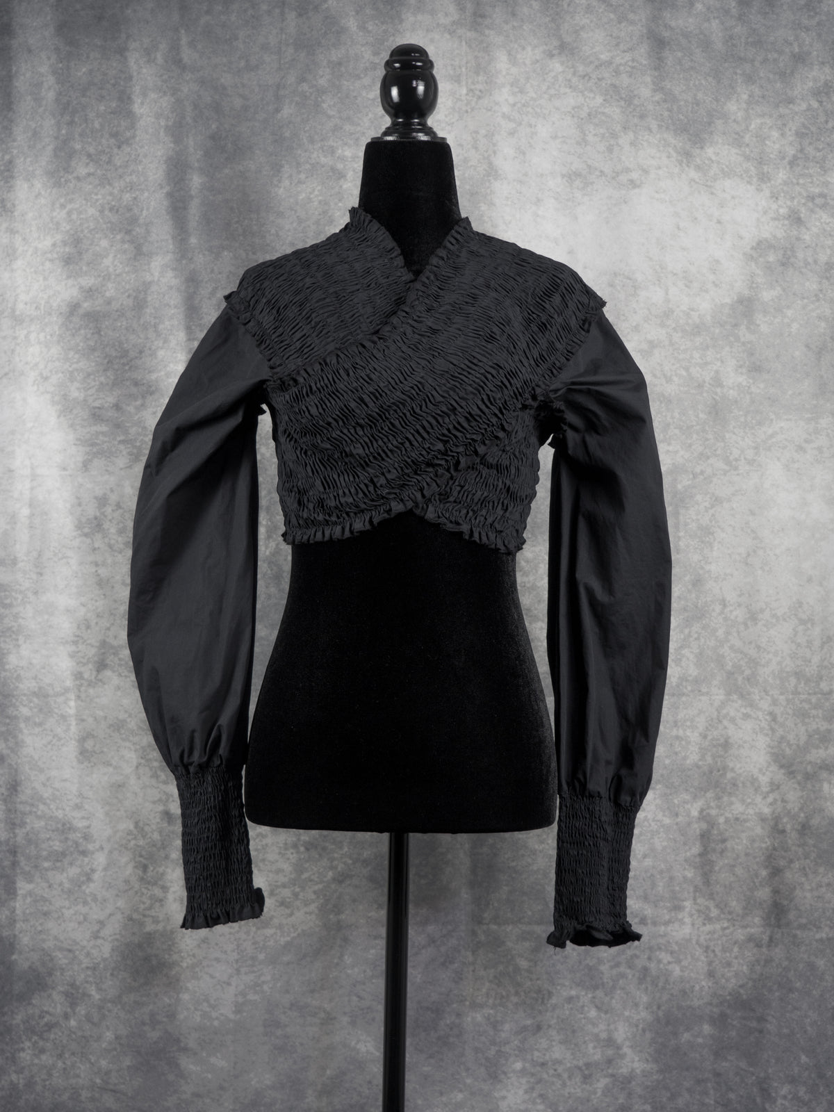A black long-sleeve blouse with a textured, crinkled wrap-style bodice and gathered sleeves, displayed on a black mannequin against a mottled gray studio background.