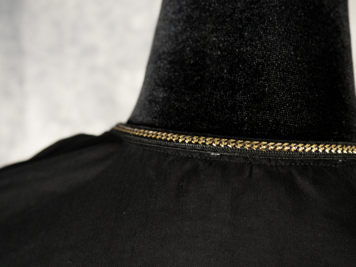 A close-up shot of the back neckline of a black dress on a mannequin. A delicate, metallic gold chain trim runs continuously along the stitched collar edge. The background features a soft, textured grey surface.