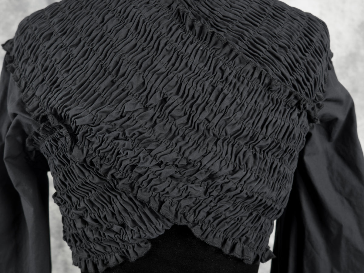 A close-up of a black long-sleeve garment with a heavily gathered, pleated smocked bodice and a ruffled hem, displayed on a mannequin against a light gray textured background.