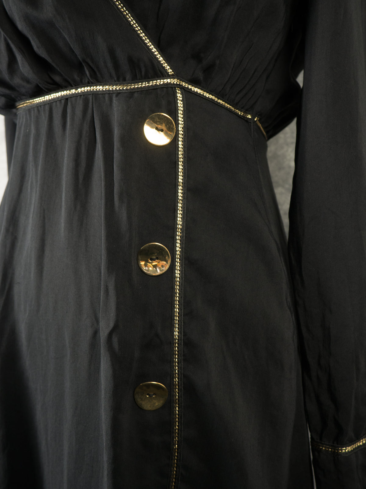 A close-up view of the waist area on a black wrap dress. It showcases a gathered golden metallic trim waistband and a crossover bodice outlined by a delicate metallic gold chain trim. Two large, shiny, round gold buttons are stitched vertically on the overlapping front panel below the waist.
