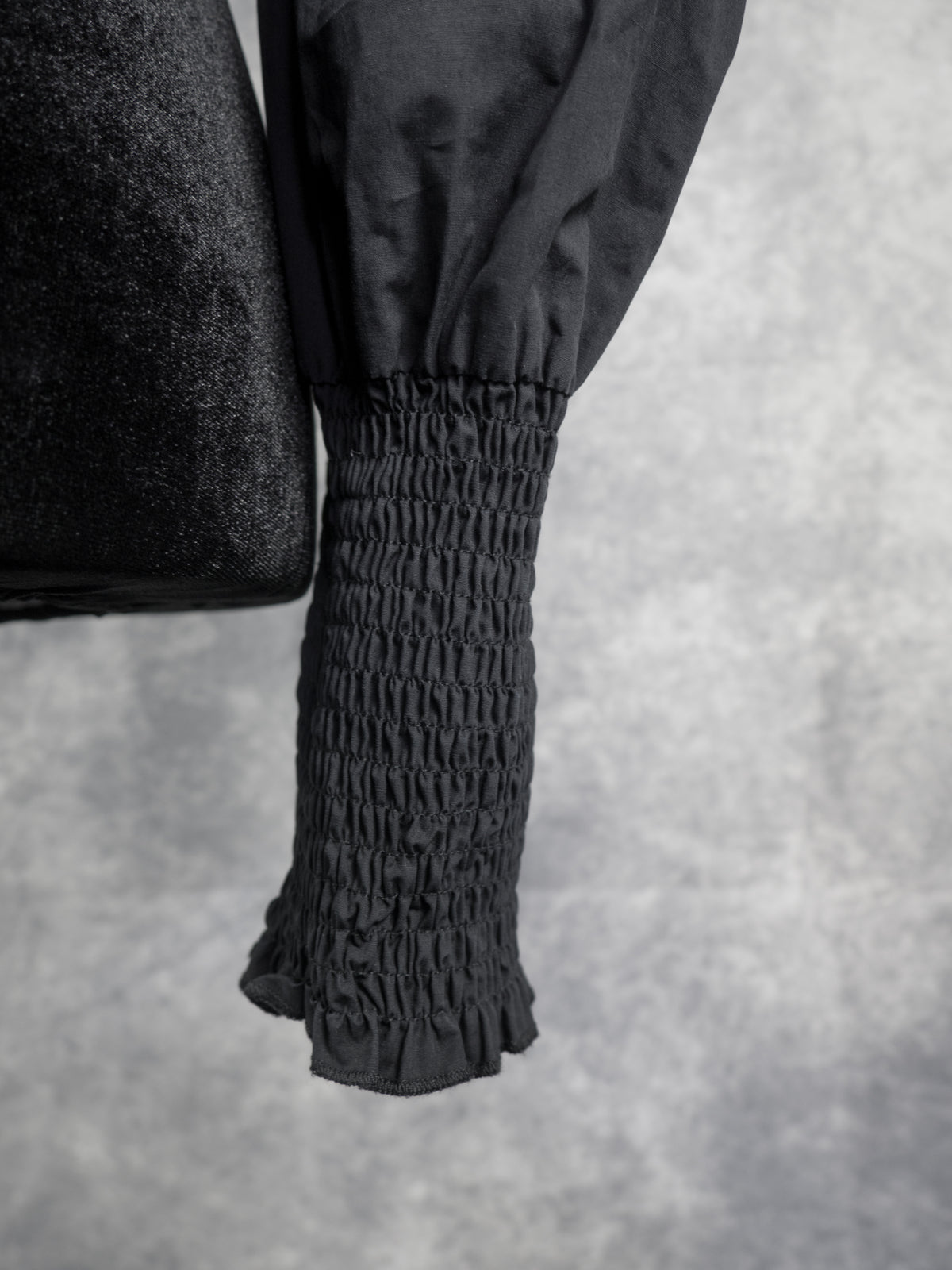 Close-up of a black garment sleeve with a gathered, smocked elastic cuff and a ruffled hem, shown against a textured gray background.