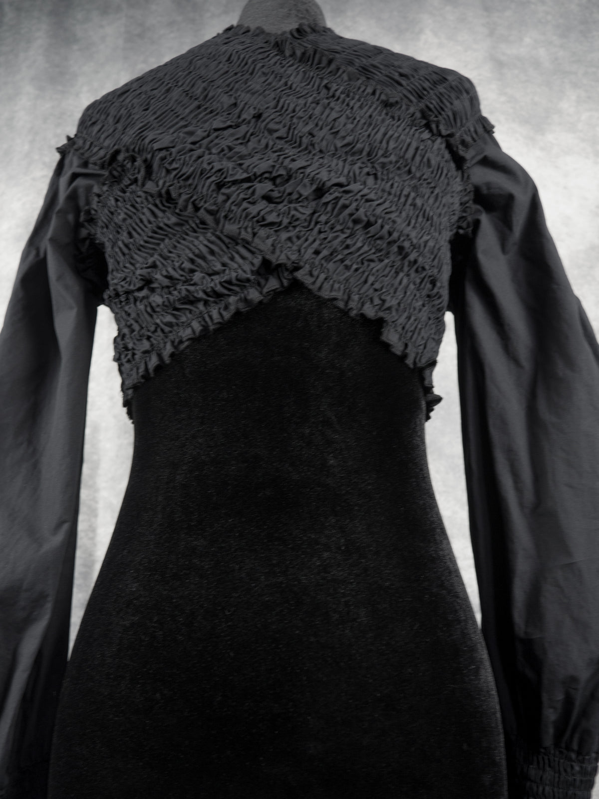 A black long-sleeve dress displayed on a mannequin, featuring a heavily textured, shimmery ruched yoke across the shoulders and upper chest, with a smoother black skirt below.