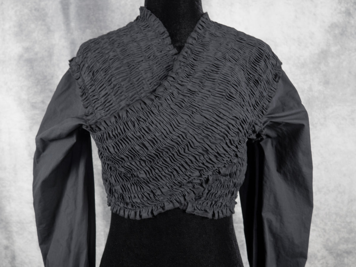 A dark gray smocked wrap-style crop top with long sleeves, displayed on a black dress form against a textured gray backdrop.