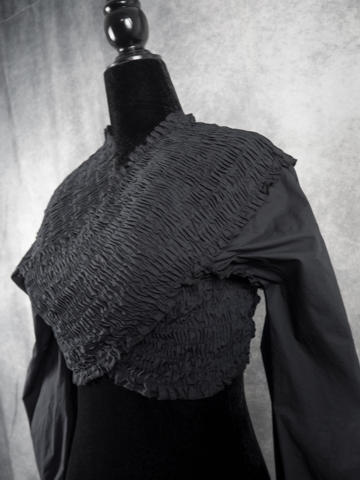 A black long-sleeve blouse with a heavily textured, ruffled smocked yoke, displayed on a black dress form mannequin against a textured gray studio backdrop.