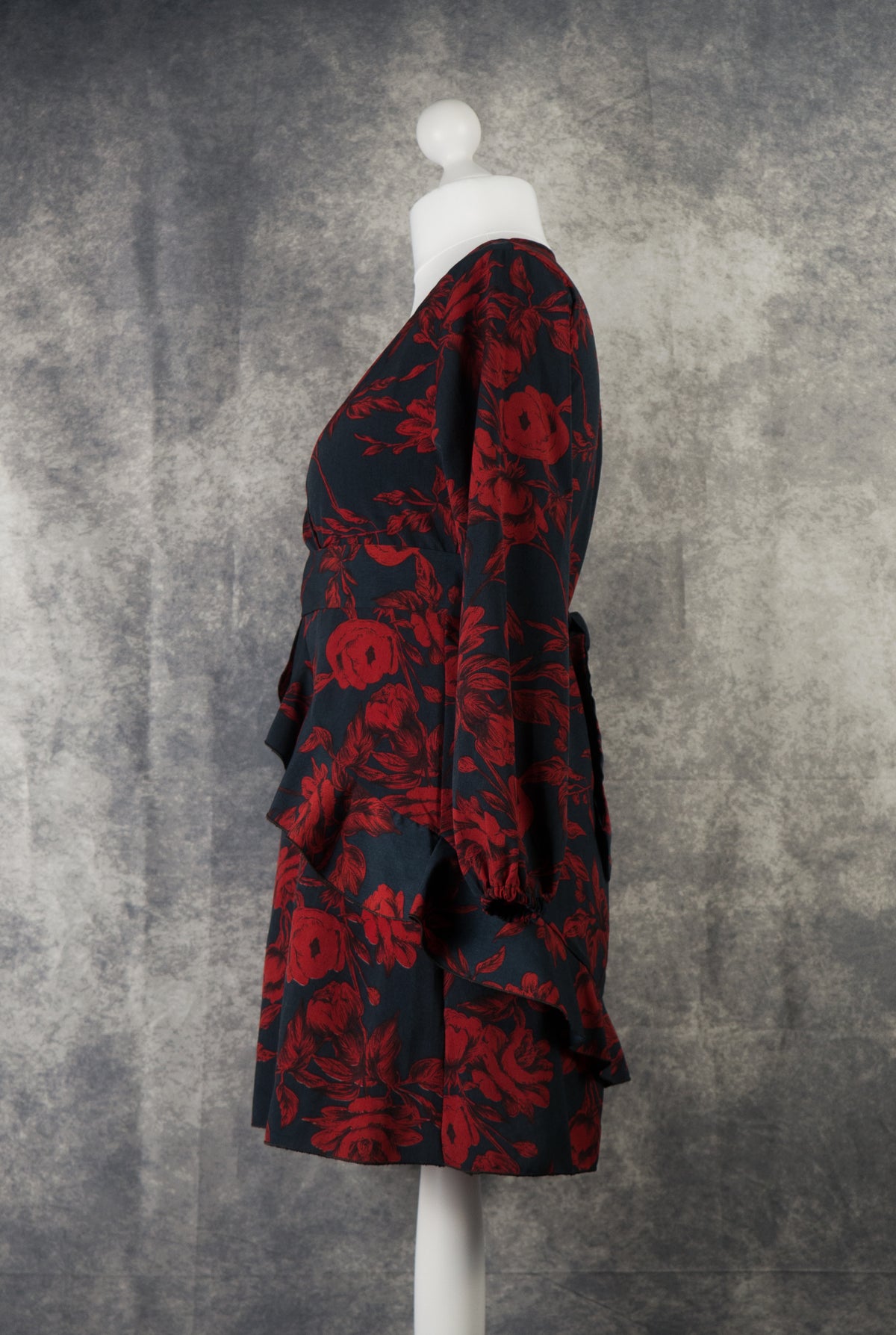A side profile view of a long-sleeved black mini dress with a dark red rose print on a white mannequin. This shot highlights the elegant drape of the long sleeve with elasticated cuffs and showcases how the layered ruffles wrap around the waist and hips. The fabric tie belt is visible at the back, set against a textured grey studio background