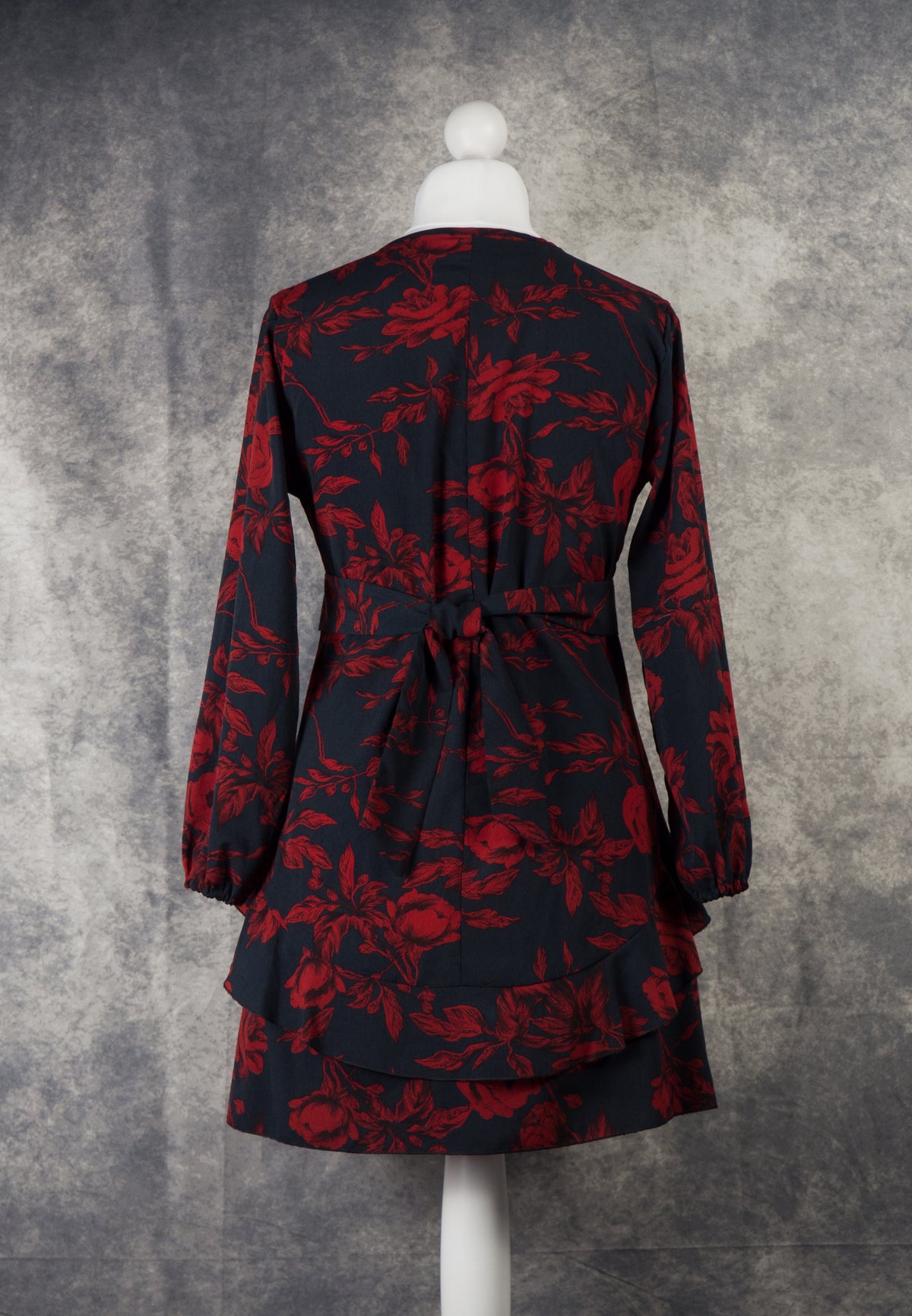 A full back view of the long-sleeved black mini dress with a dark red rose print on a white mannequin. It features a high neckline and highlights a matching fabric tie belt neatly knotted at the center of the waist. The hemline shows a tiered ruffle layer across the back, completing the look against a textured grey studio background.