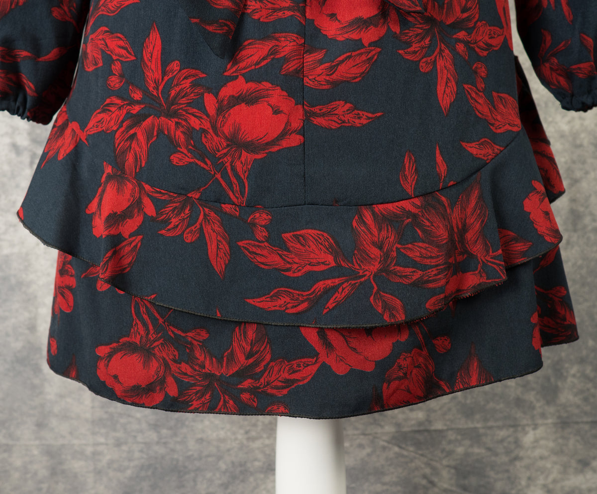 Close-up detail of the lower back portion of a black mini dress featuring a dark red rose print. The shot highlights the tiered, horizontal ruffle layer running across the back hem, emphasizing the volume and layered design of the skirt against a textured grey background.