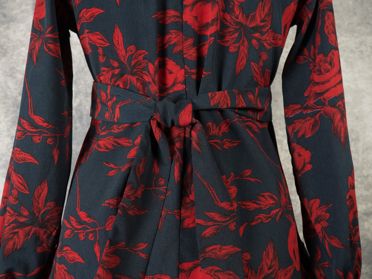 A close-up view of the back of a black dress with a dark red rose print, focusing on the waistline. A matching fabric tie belt is neatly tied in a secure knot at the center, highlighting the tailored waist and the continuous floral and leaf pattern against a textured grey background.