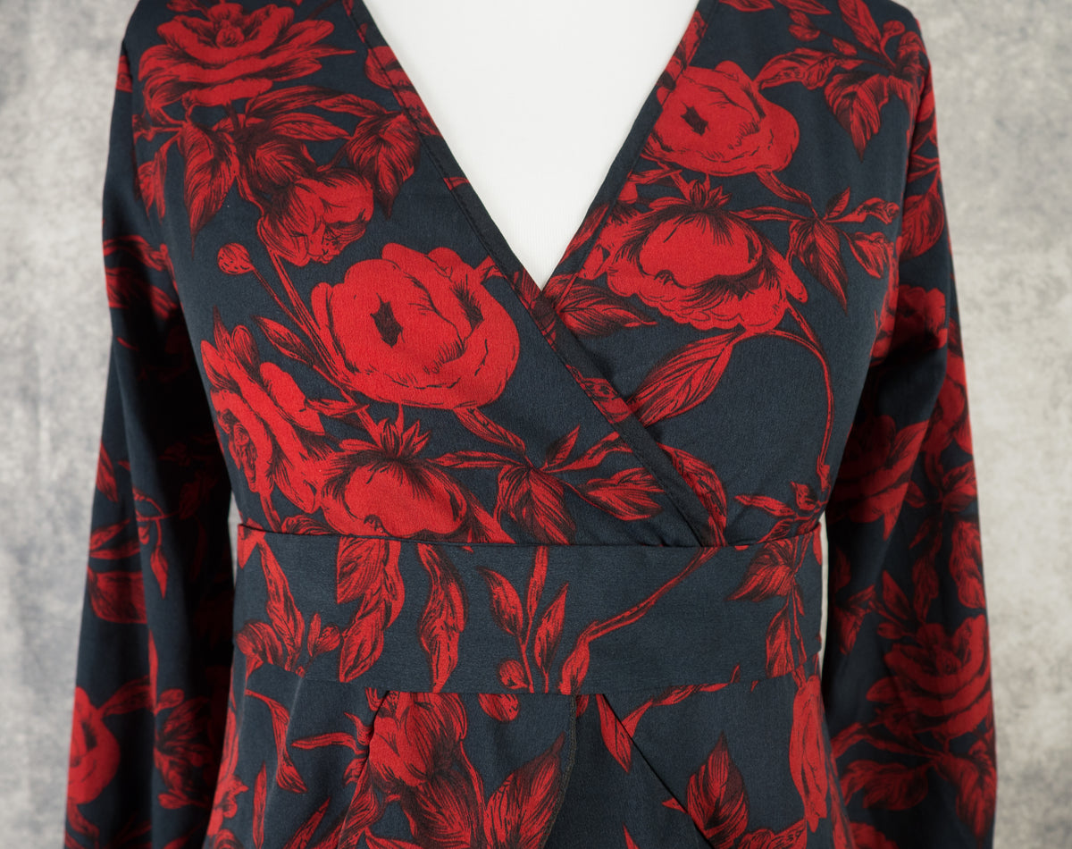 Close-up detail of the front bodice of a black dress with a dark red rose print on a mannequin. The shot highlights the deep faux-wrap V-neckline and the structured waistband panel right below the bust, showcasing the vibrant floral pattern against a textured grey background.