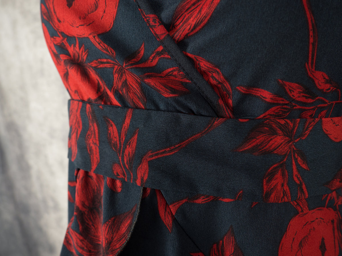 An angled close-up shot of the front bodice of a black dress with a dark red rose pattern. The image emphasizes the wrap-style overlapping fabric at the V-neckline and the flat waistband panel below the bust, highlighting the smooth texture and floral details against a textured grey background.