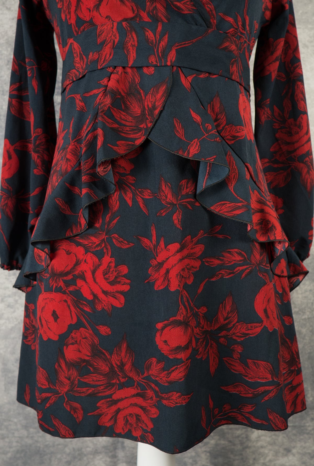 A front close-up view of the skirt on a black dress with a dark red rose print. The shot highlights the structured waist panel and the dramatic, asymmetrical cascading ruffles that form a layered design down the front of the mini-length skirt, shown against a grey background.