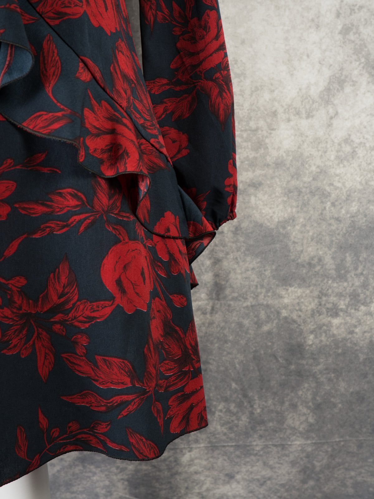 A detailed side view of a black mini dress featuring a rich red rose pattern. The image showcases the structured, cascading ruffle trim on the skirt alongside a long sleeve with a gathered elastic cuff. The contrast between the vibrant floral print and the textured grey background highlights the garment's design.