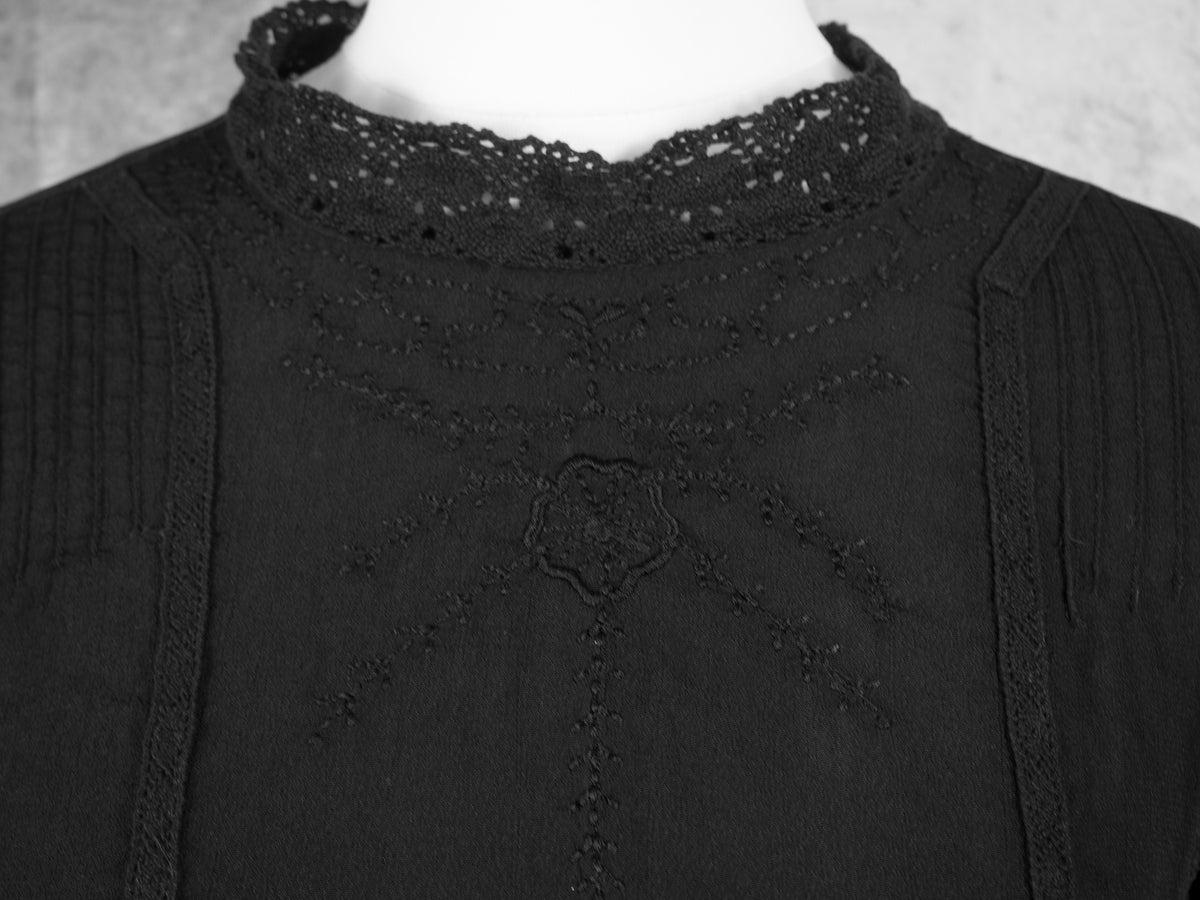 A close-up view of the hemline of a black dress, highlighting a subtle embroidered horizontal lace trim running just above the bottom edge. The lightweight, slightly sheer quality of the black fabric is visible along the fluid hemline against a soft grey background.