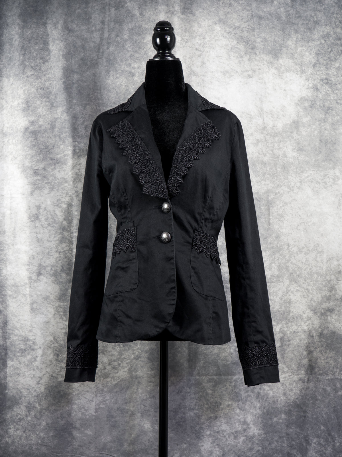  A tailored black alternative blazer displayed on a black female mannequin against a textured grey studio background. The jacket features notched lapels adorned with intricate black floral lace trim along the edges. It secures with two ornate silver-tone statement buttons at the waist, complemented by matching lace details across the side pockets and cuffs to create a gothic, structured silhouette.