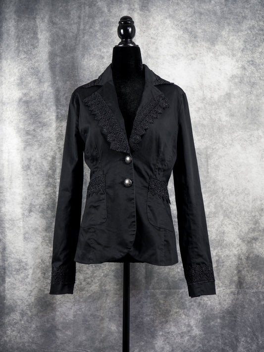  A tailored black alternative blazer displayed on a black female mannequin against a textured grey studio background. The jacket features notched lapels adorned with intricate black floral lace trim along the edges. It secures with two ornate silver-tone statement buttons at the waist, complemented by matching lace details across the side pockets and cuffs to create a gothic, structured silhouette.