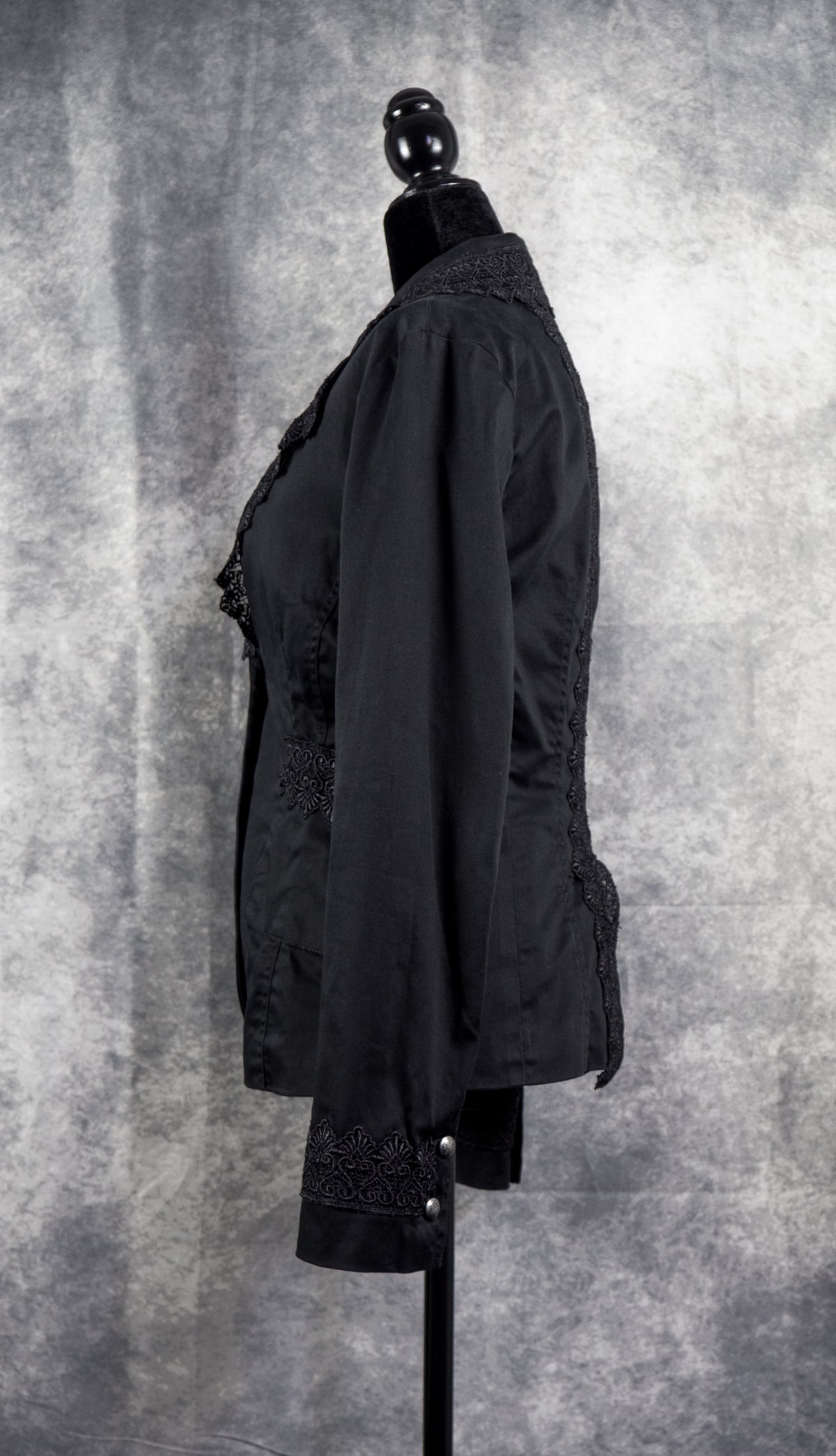  A tailored black alternative blazer displayed on a black female mannequin against a textured grey studio background. The jacket features notched lapels adorned with intricate black floral lace trim along the edges. It secures with two ornate silver-tone statement buttons at the waist, complemented by matching lace details across the side pockets and cuffs to create a gothic, structured silhouette.