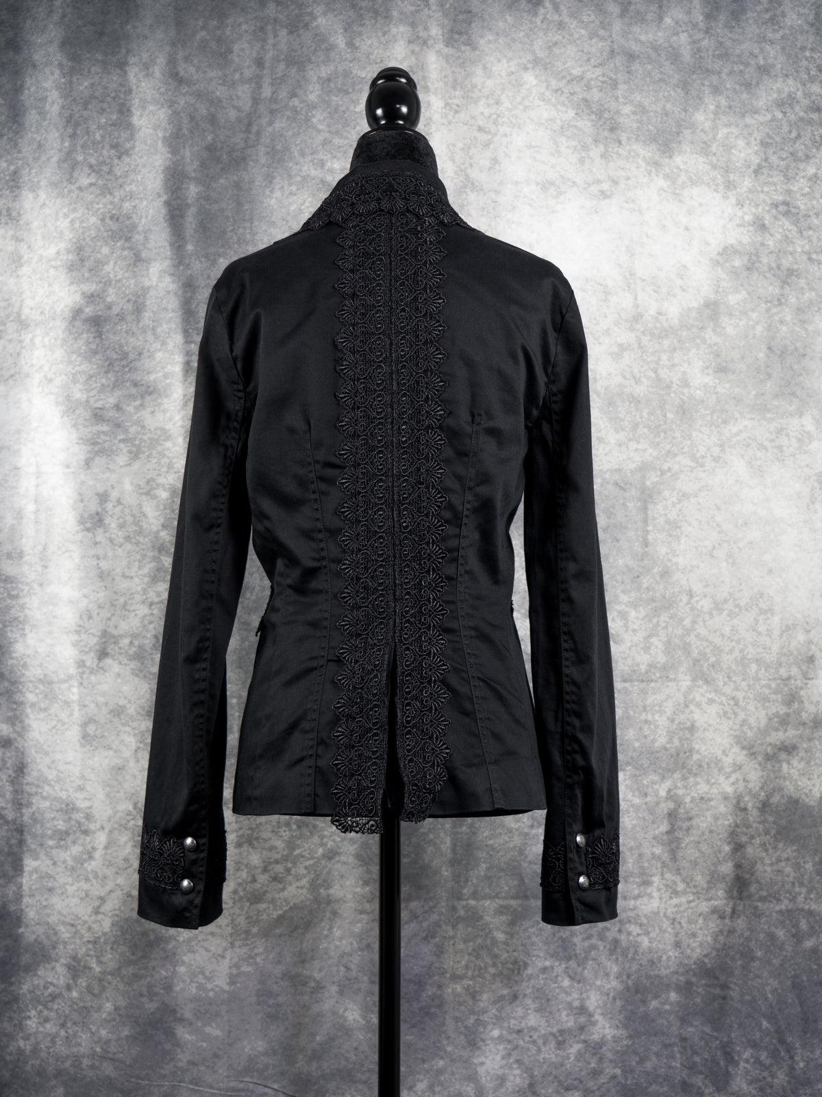  A tailored black alternative blazer displayed on a black female mannequin against a textured grey studio background. The jacket features notched lapels adorned with intricate black floral lace trim along the edges. It secures with two ornate silver-tone statement buttons at the waist, complemented by matching lace details across the side pockets and cuffs to create a gothic, structured silhouette.