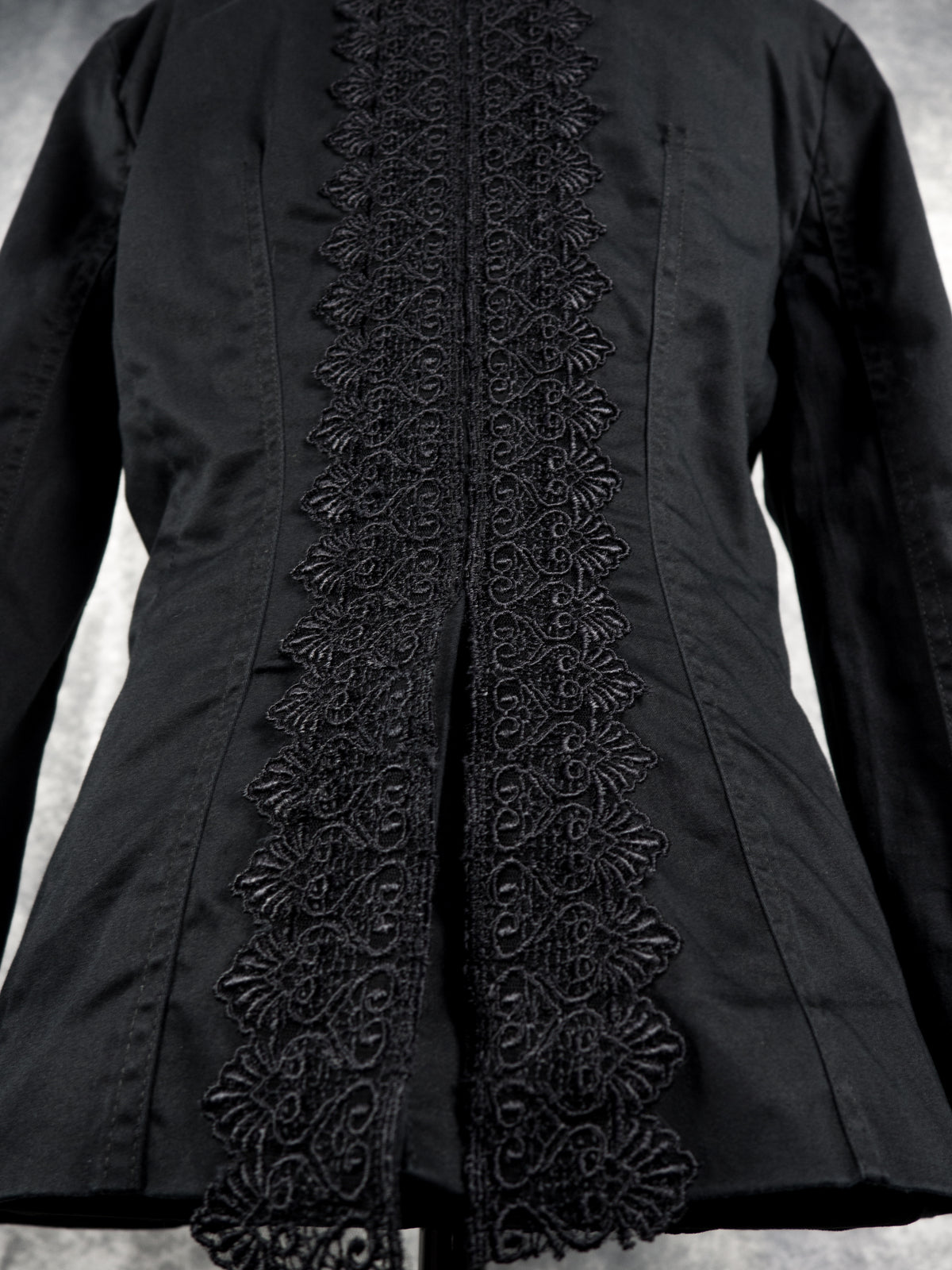 A close-up of a black long-sleeve jacket featuring a prominent vertical panel of intricate black floral lace embroidery down the center front.
