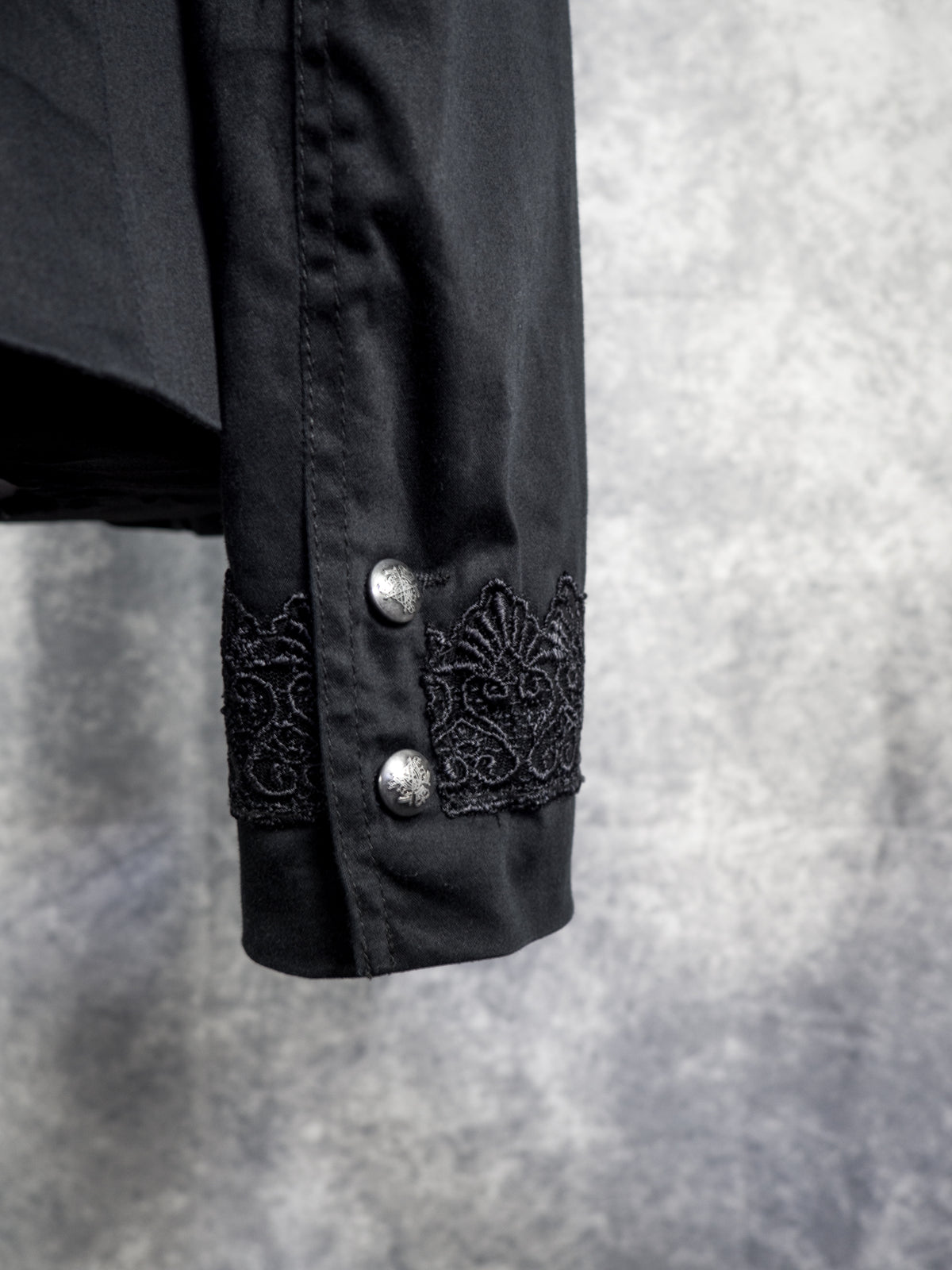 Close-up of a black long-sleeve blazer sleeve with ornate black lace trim and two silver-toned buttons on the cuff, set against a textured gray background.