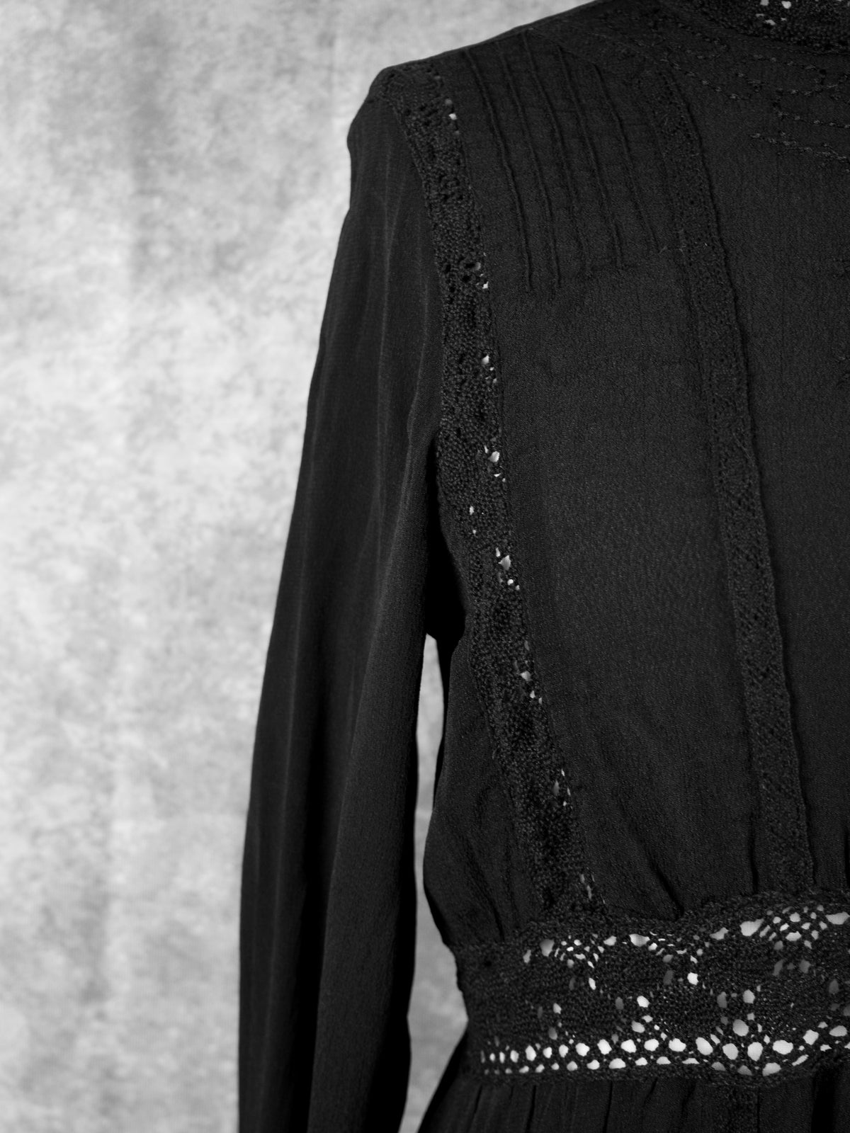 A close-up angled shot of the front bodice on a black dress, highlighting a vertical lace insert running alongside a pintuck pleated panel near the shoulder. The openwork geometric lace waistband is visible below, contrasting with the soft, flowing fabric of the sleeve against a textured grey background.