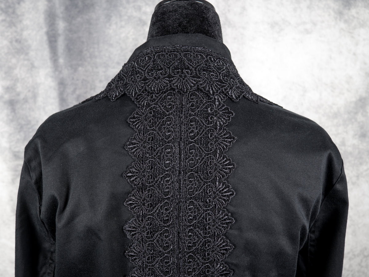 A close-up view from behind focusing on the upper back and collar of the black blazer. A wide, ornate panel of black lace featuring symmetric, swirling heart and fan-shaped motifs runs vertically down the central spine of the jacket. The high, structured collar stands upright above the shoulders, bordered by the same intricate lace trim pointing outward.
