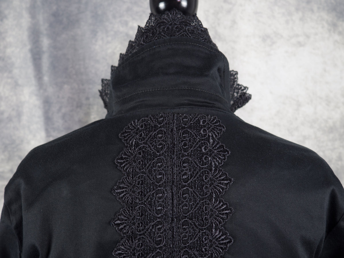 A close-up view from behind focusing on the upper back and collar of the black blazer. A wide, ornate panel of black lace featuring symmetric, swirling heart and fan-shaped motifs runs vertically down the central spine of the jacket. The high, structured collar stands upright above the shoulders, bordered by the same intricate lace trim pointing outward.