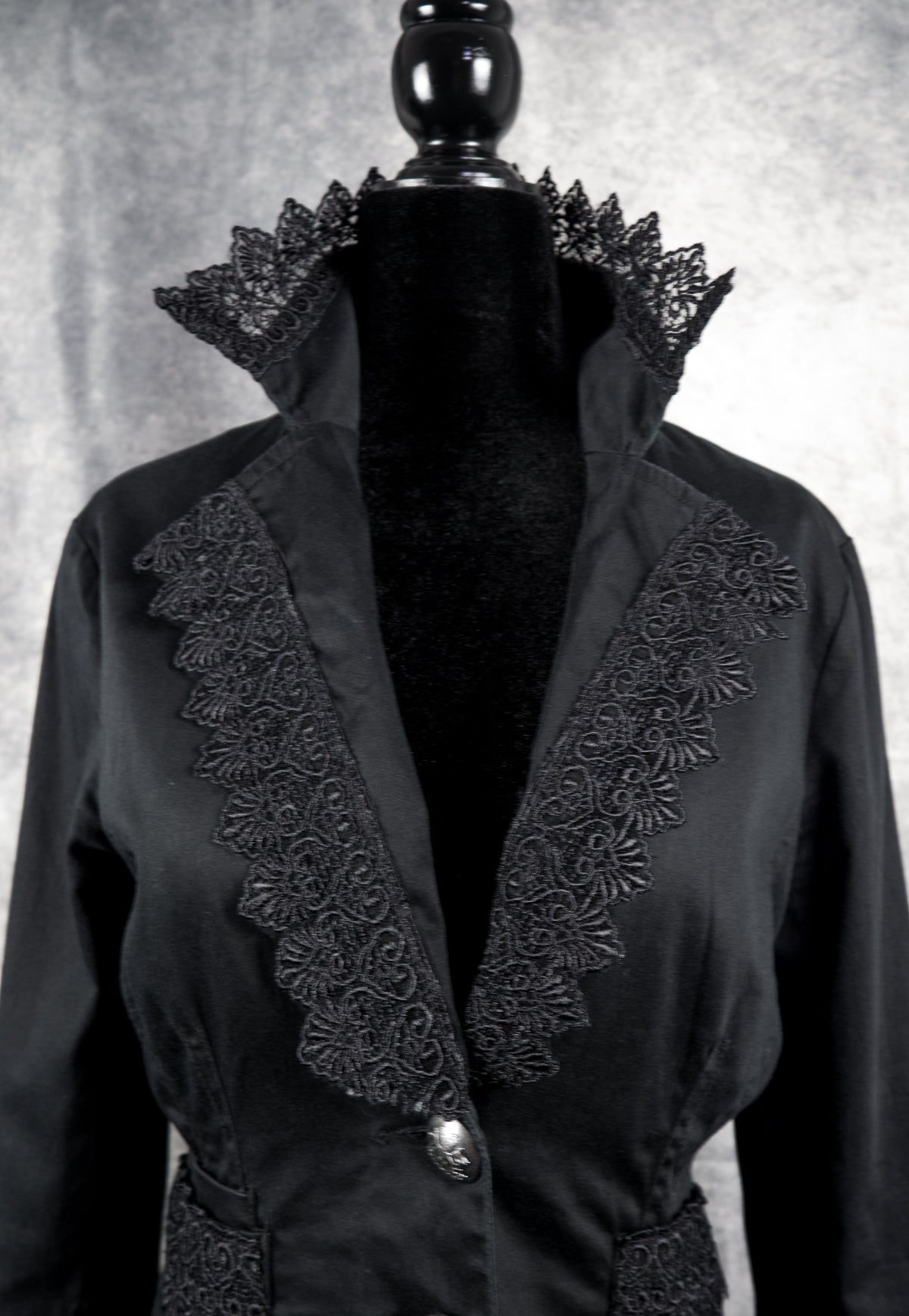 A black long-sleeve jacket with dramatic ruffled lace trim and a single button closure, displayed on a black mannequin torso against a textured gray background.