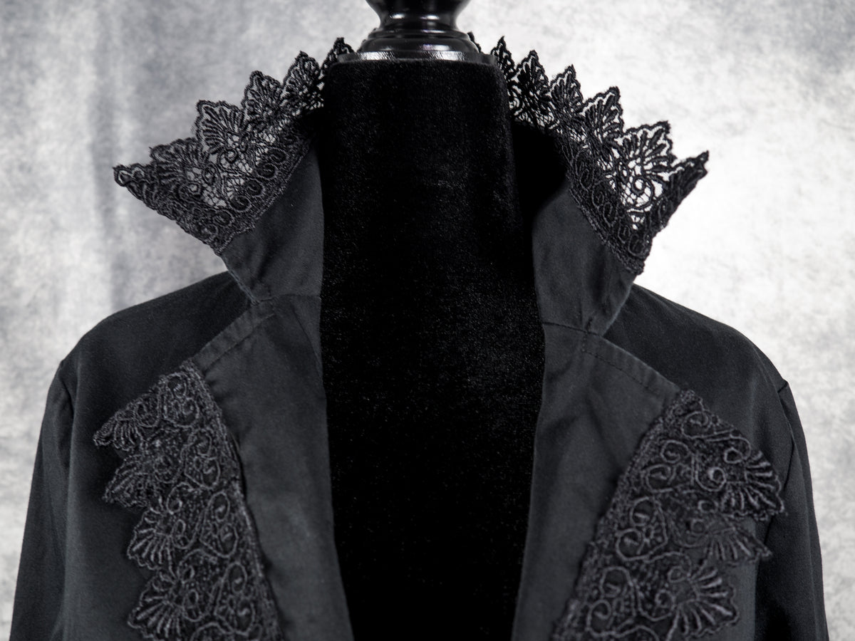 A close-up view focusing on the high collar and lapels of the black gothic blazer on the mannequin. The collar stands upright, framed with a flared border of intricate, fan-shaped black floral lace that stands out against the grey background. The matching lace continues down the edges of the open V-neckline lapels, highlighting the textured and detailed craftsmanship of the garment.