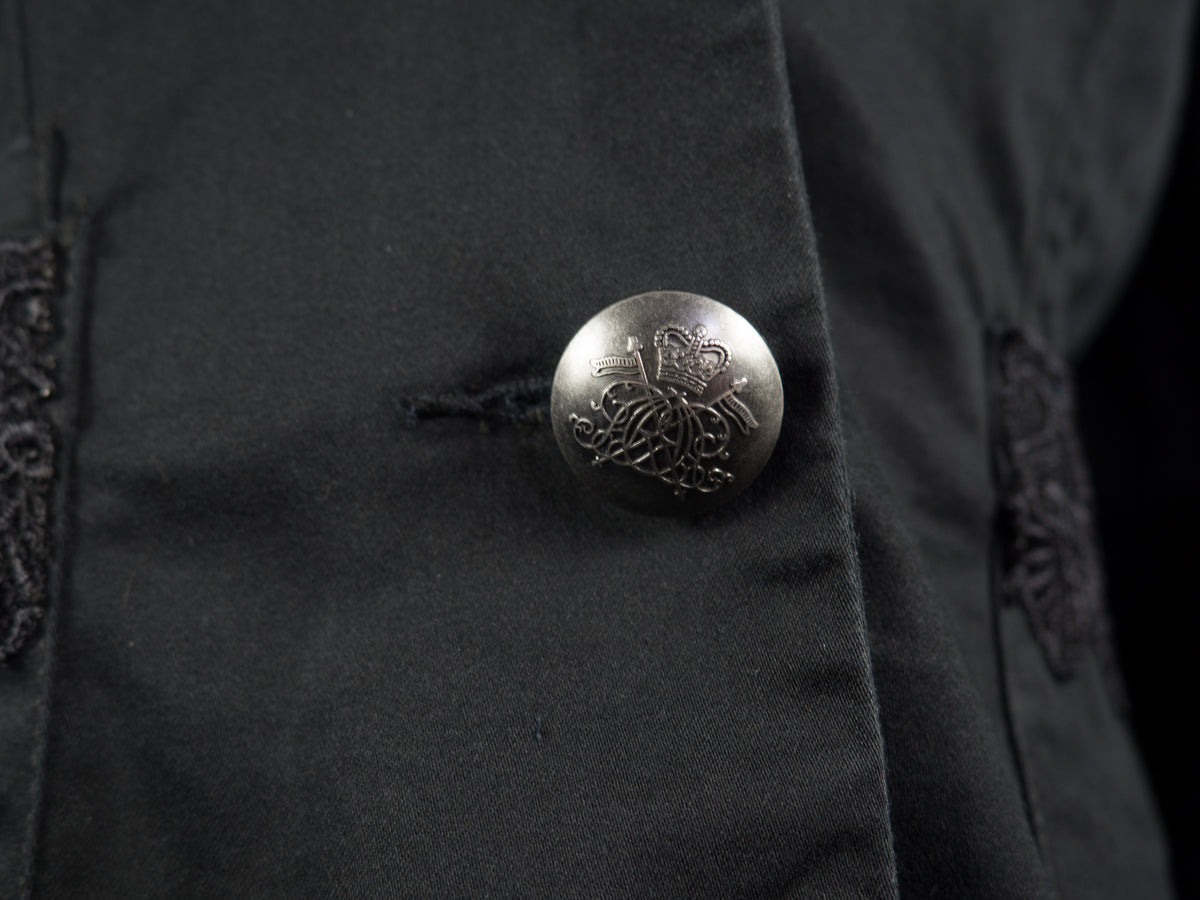Close-up of a black garment featuring a prominent round silver metal button with an embossed floral and crown motif.