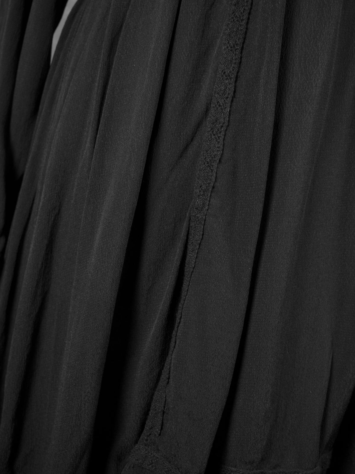 A detailed close-up of the black dress skirt, showcasing the soft gathers and folds of the lightweight crêpe fabric. A vertical delicate embroidered lace trim runs down the front, highlighting the flowing texture and bohemian design of the piece.
