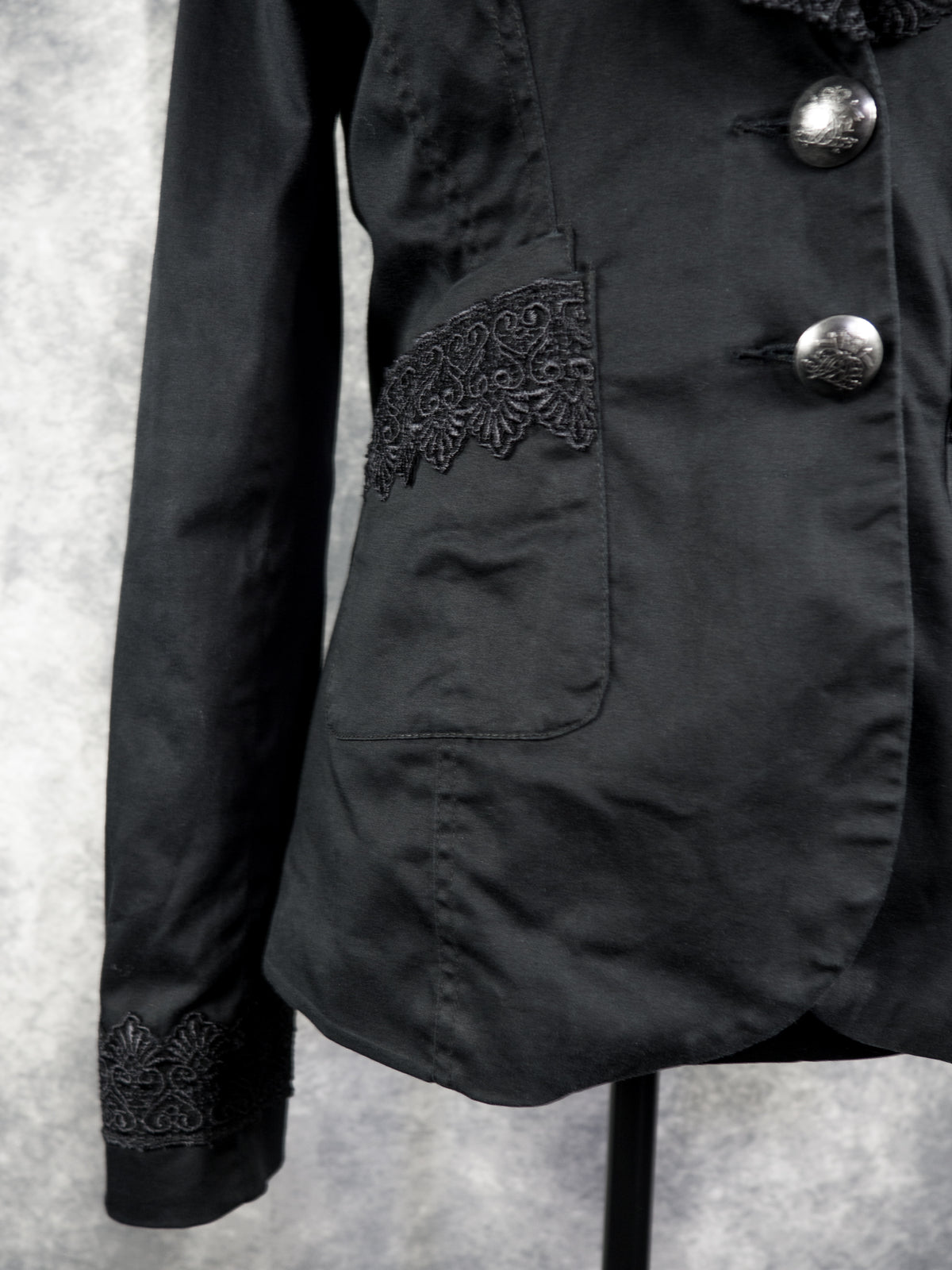 A close-up view focusing on the lower right section of the black blazer. It shows a patch pocket adorned with a wide band of intricate black lace trim running horizontally along its top opening. To the right, two ornate silver-tone statement buttons are fastened at the waist, while the bottom of the sleeve cuff is visible in the lower left corner, featuring matching decorative lace details.