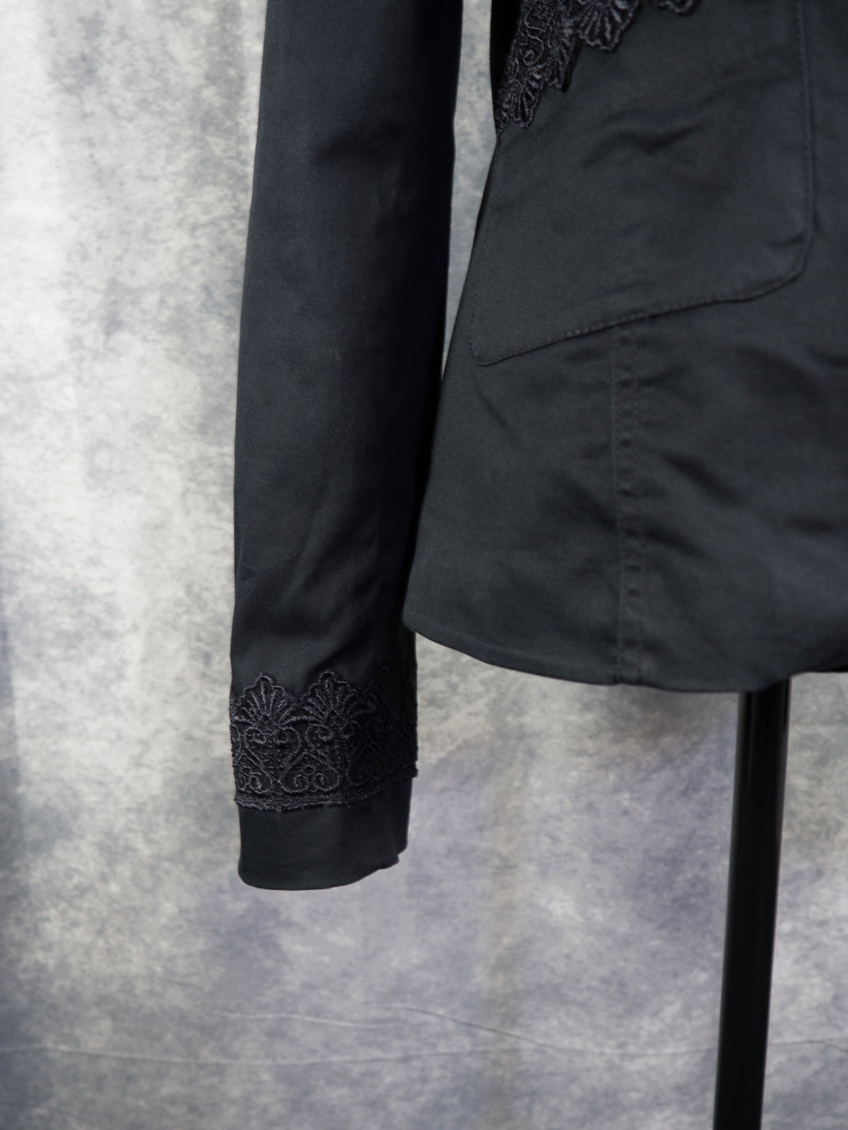 A close-up view focusing on the lower sleeve and cuff of the black blazer. The wrist cuff is decorated with a wide band of intricate black lace trim featuring symmetric, fan-shaped motifs. To the right, a portion of the blazer's clean, structured side hem is visible against the textured grey background.