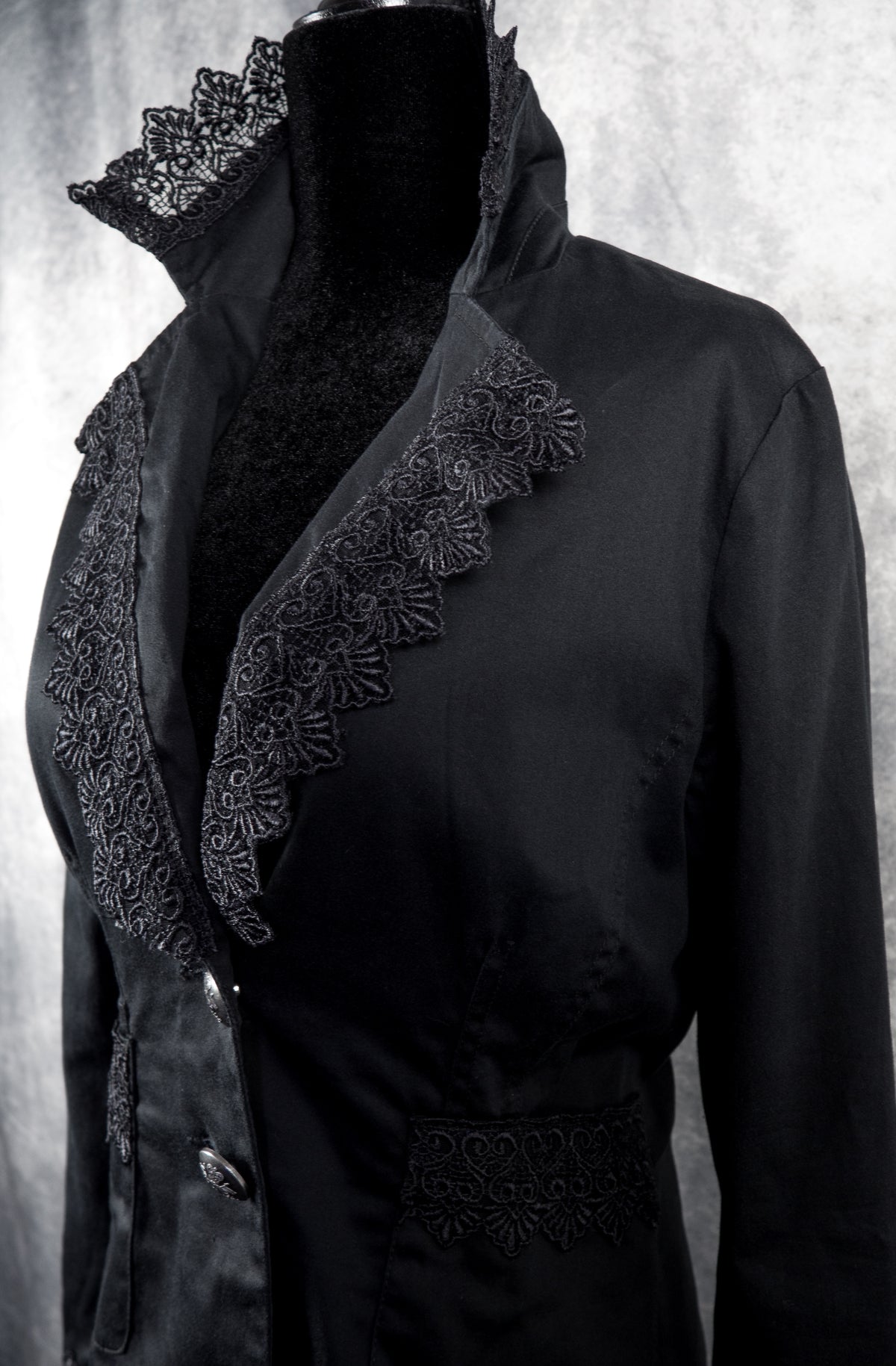 A close-up, three-quarter side view of the black gothic blazer on the mannequin. The shot highlights the structured line of the open lapels and the dramatic high collar, both heavily bordered with ornate black floral lace. The two silver statement buttons are visible at the front closure, along with the lace trim detailed across the left side pocket.